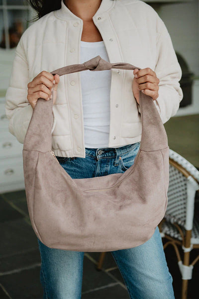A woman in a cream jacket, white top, and jeans holds the Katydid Khaki Lightweight Suede Tote Bag with both hands. The photo, cropped above her nose, highlights the stylish bag and her chic outfit.