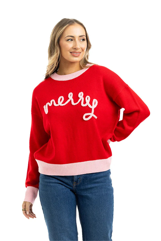 A woman models the Katydid Merry Embroidered Sweater Christmas Woman, featuring merry in white script, a light pink collar and cuffs, and blue jeans. She stands smiling with one hand on her hip against a white backdrop.