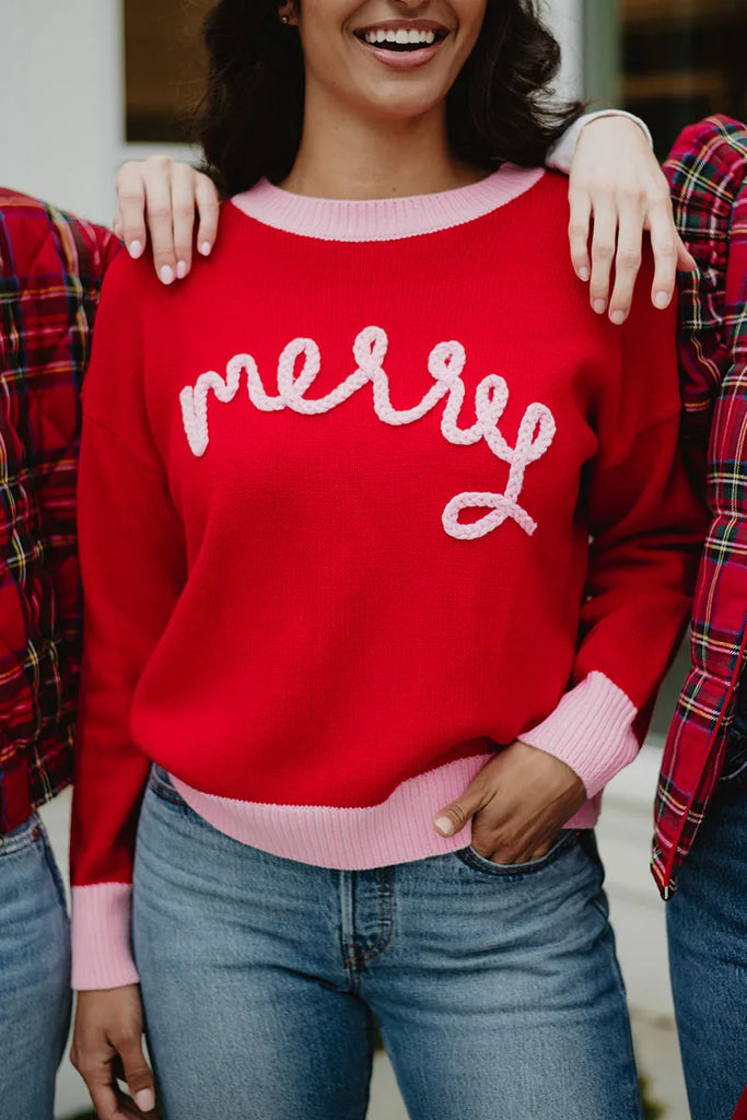 Three people stand side by side. The center person wears the Katydid Merry Embroidered Sweater Christmas Woman—a red sweater with “merry” in white, light pink cuffs and collar, and blue jeans. Only one smiling face and their torsos are visible.