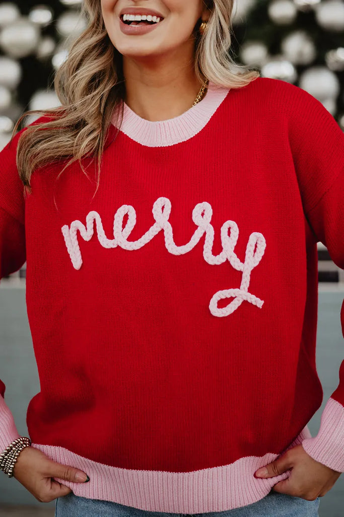 A woman smiles in a Katydid Merry Embroidered Sweater Christmas Woman—a red knit with merry in pink yarn across the front, plus light pink cuffs and collar. The blurred background features festive lights.