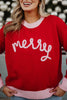 A woman smiles in a Katydid Merry Embroidered Sweater Christmas Woman—a red knit with merry in pink yarn across the front, plus light pink cuffs and collar. The blurred background features festive lights.