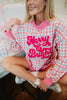 A woman sits cross-legged on a chair, smiling in the Katydid Merry  Bright Plaid Mockneck Sweatshirt—a festive women’s holiday pullover in pink, blue, and white. Gift boxes wrapped in white paper and pink ribbon are nearby.