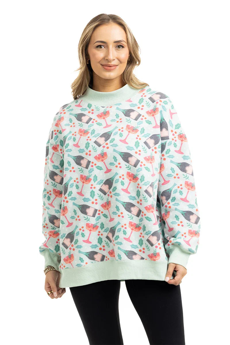 A person wears the Katydid Holiday Champagne, Coupes  Holly Mockneck Sweatshirt, standing with hands relaxed by their sides and smiling at the camera against a white background.
