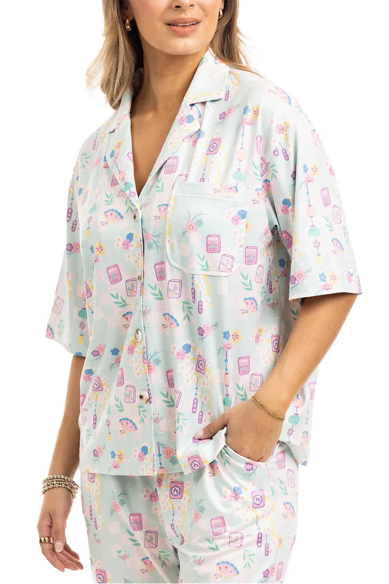 A woman models the Katydid Mahjong Pieces Pajama Pants Set, a light-colored, short-sleeve collared set with buttons and a playful pastel Mahjong pattern. She accessorizes with bracelets, adding charm to these cute pajamas.
