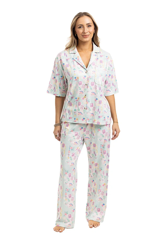 A woman smiles barefoot in the Katydid Mahjong Pieces Pajama Pants Set, featuring a light, loose fit and playful pattern. The short-sleeved button-up top and long pants create a stylish look against a plain white background.