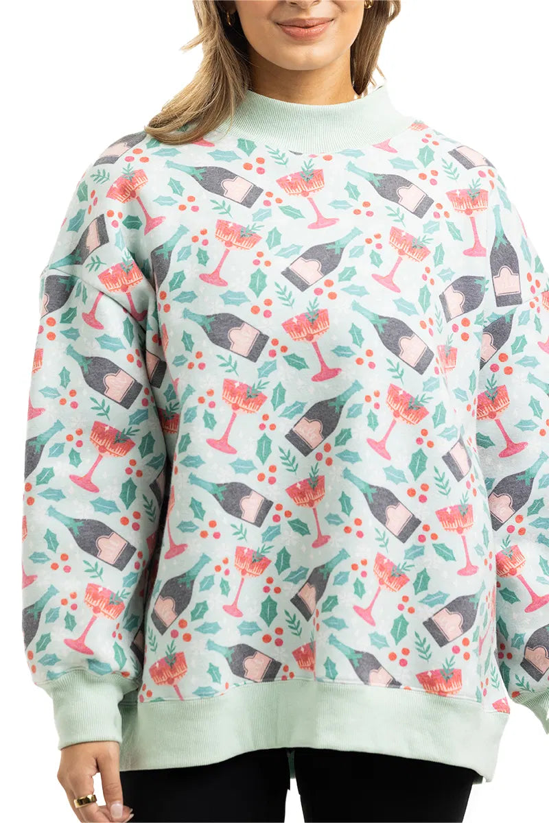 A woman wears the Katydid Holiday Champagne, Coupes  Holly Mockneck Sweatshirt, featuring a festive print of wine bottles, coupes, holly leaves, and red berries. Only her upper body is shown as she smiles in this cozy light green top.