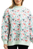A woman wears the Katydid Holiday Champagne, Coupes  Holly Mockneck Sweatshirt, featuring a festive print of wine bottles, coupes, holly leaves, and red berries. Only her upper body is shown as she smiles in this cozy light green top.