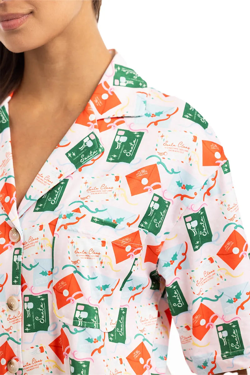 A woman wears Katydids Santas Mail Christmas Pajamas for Women—a button-up shirt with green and red postage stamps, dolphins, sunglasses, and starfish on white—perfect to pair with holiday pajama pants. Only her lower face is shown.
