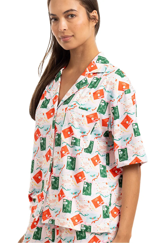 A woman in Katydids Santas Mail Christmas Pajamas for Women, featuring a short-sleeved button-up top with colorful green and orange carton prints on white, smiles softly—ideal comfy Christmas sleepwear.