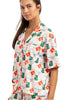 A woman in Katydids Santas Mail Christmas Pajamas for Women, featuring a short-sleeved button-up top with colorful green and orange carton prints on white, smiles softly—ideal comfy Christmas sleepwear.