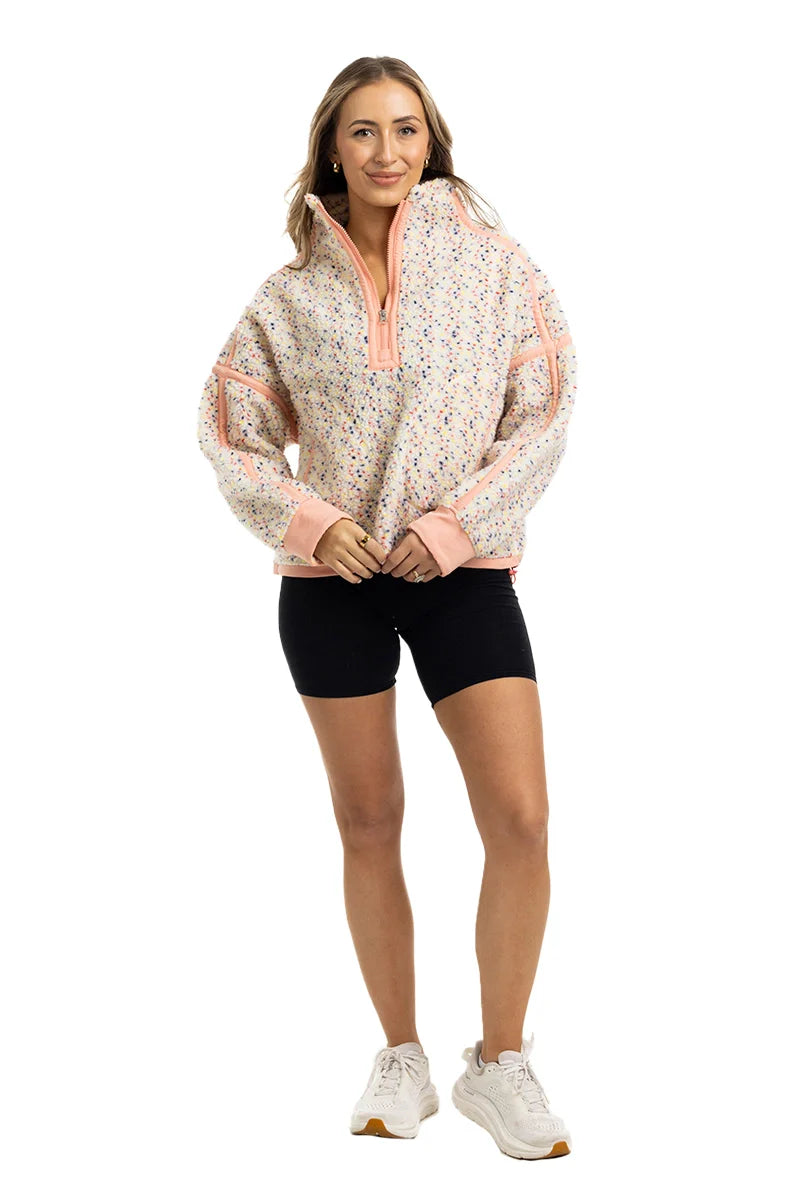 A woman with long hair smiles in a Katydid Confetti Print Womens Pullover with peach accents, paired with black shorts and white sneakers, posing against a white background.