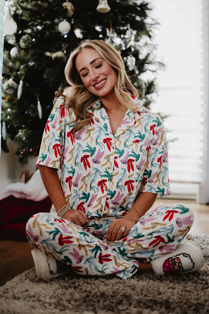 A smiling woman sits cross-legged in front of a decorated Christmas tree, wearing the Katydid Whimsical Bow Pajamas Set and slippers, with her hands resting comfortably on her knees.