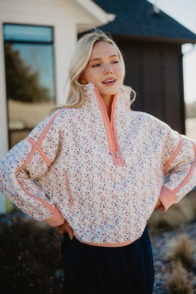 A woman smiles with her hands on her hips outside a house, wearing the Katydid Confetti Print Womens Pullover featuring a multicolored confetti design and peach accents.