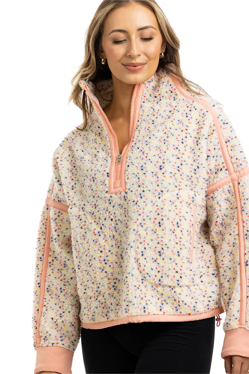 A woman wears the Katydid Confetti Print Womens Pullover, featuring a half-zip front, peach trim, and playful speckled fleece. She smiles softly with her eyes closed, her long light brown hair framing her face.