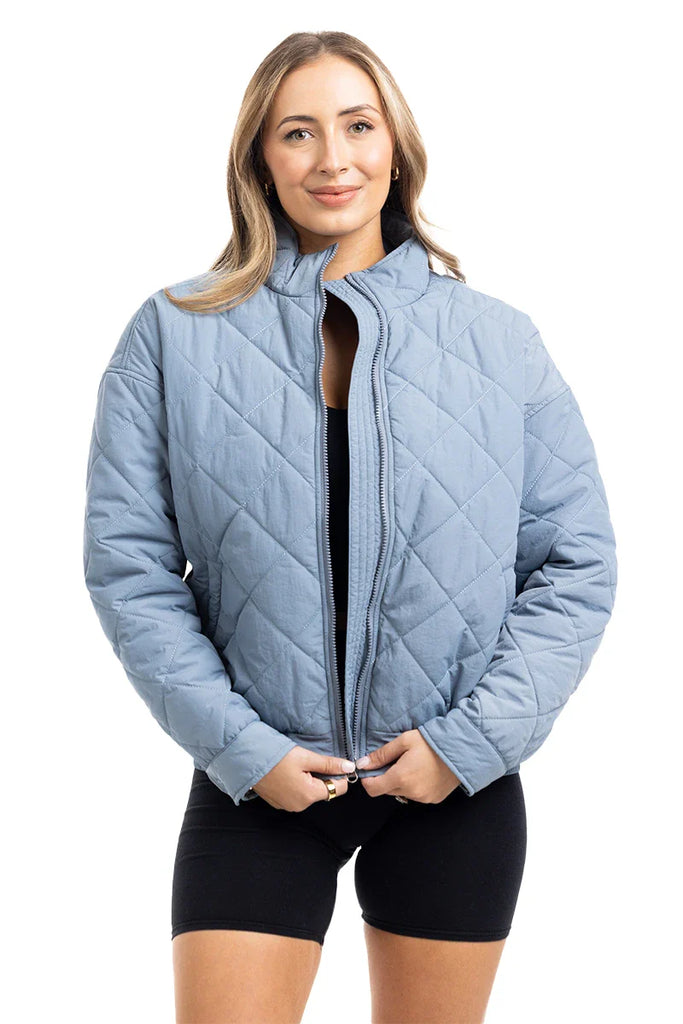 A woman with light brown hair faces forward, smiling slightly as she holds the zipper of the Katydid Steel Blue Sporty Diamond Quilted Womens Jacket over a black outfit.