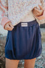 A person in a speckled jacket and white top lifts the hem to reveal Katydid Navy Bubble Skorts for Women, featuring an elastic waistband.