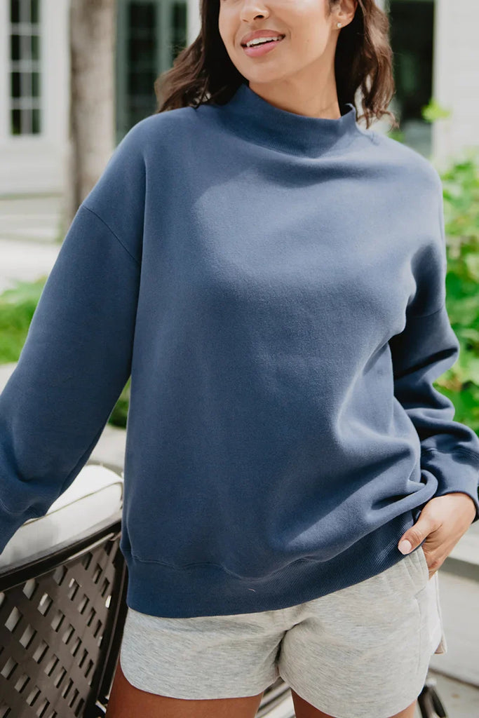 A woman wears the Katydid Navy Mockneck Sweatshirt and light gray shorts, standing outdoors with a hand on a chair, greenery and a building softly blurred in the background.