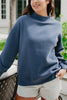 A woman wears the Katydid Navy Mockneck Sweatshirt and light gray shorts, standing outdoors with a hand on a chair, greenery and a building softly blurred in the background.