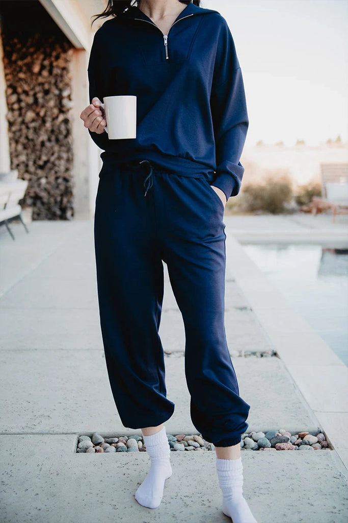 A person stands by a pool in the Katydid Classic Navy Two-Piece Pants Set—long-sleeve zip-up top and jogger pants, hands in pockets, holding a white mug and wearing white socks. The head is not visible.