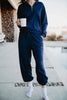 A person stands by a pool in the Katydid Classic Navy Two-Piece Pants Set—long-sleeve zip-up top and jogger pants, hands in pockets, holding a white mug and wearing white socks. The head is not visible.