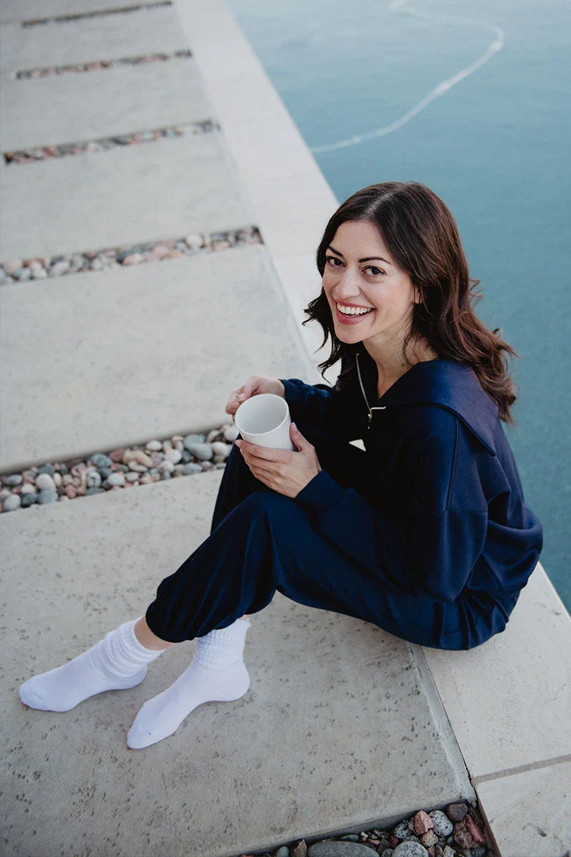 A woman with brown hair, dressed in the Katydid Classic Navy Two-Piece Pants Set and white socks, sits on a concrete edge by the water, smiling at the camera while holding a white mug. Pebbles are scattered along the walkway beside her.
