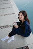 A woman with brown hair, dressed in the Katydid Classic Navy Two-Piece Pants Set and white socks, sits on a concrete edge by the water, smiling at the camera while holding a white mug. Pebbles are scattered along the walkway beside her.