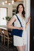 A woman in a sleeveless white dress stands indoors, smiling and holding a door frame. She carries the Katydid Navy Woven Everywhere Small Crossbody with an adjustable strap. A dining table and wooden chairs are visible behind her.