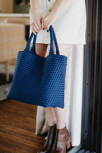 A person in a cream dress and brown ankle boots holds the Katydid Navy Lightweight Woven Tote by the handles while standing indoors near a doorway.