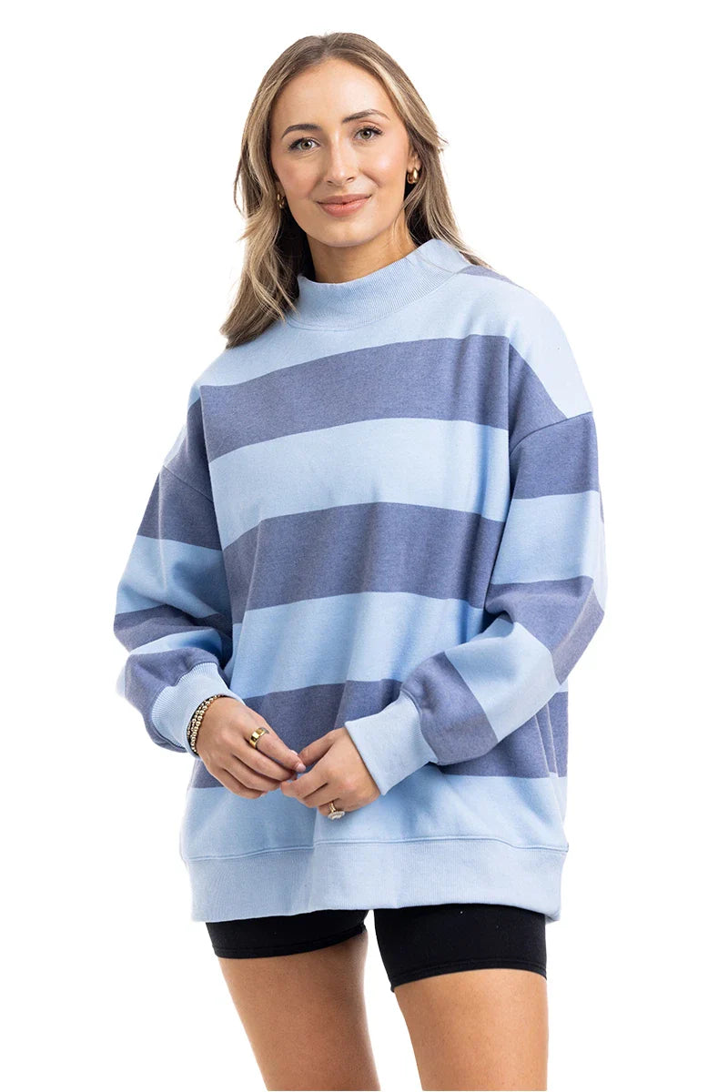 A woman with light brown hair wears the Katydid Light Blue and Navy Striped Mockneck Sweatshirt with black shorts, smiling against a white background—an ideal choice for effortless fall layering.