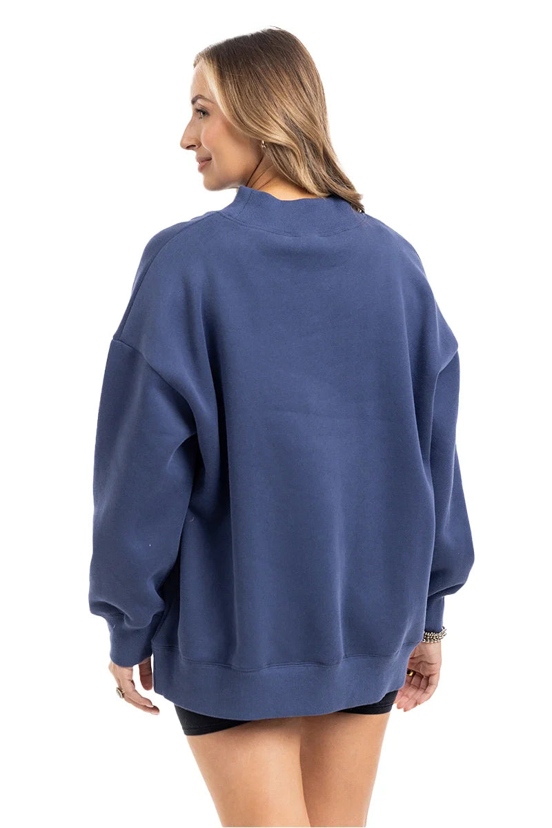 A woman with light brown hair models the Katydid Navy Mockneck Sweatshirt for women, paired with black shorts. She stands smiling, shown from the back against a white background.