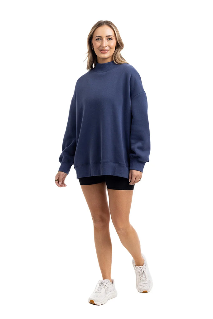 A woman smiles in a Katydid Navy Mockneck Sweatshirt, paired with black shorts and white sneakers, standing against a plain white background.