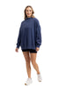 A woman smiles in a Katydid Navy Mockneck Sweatshirt, paired with black shorts and white sneakers, standing against a plain white background.