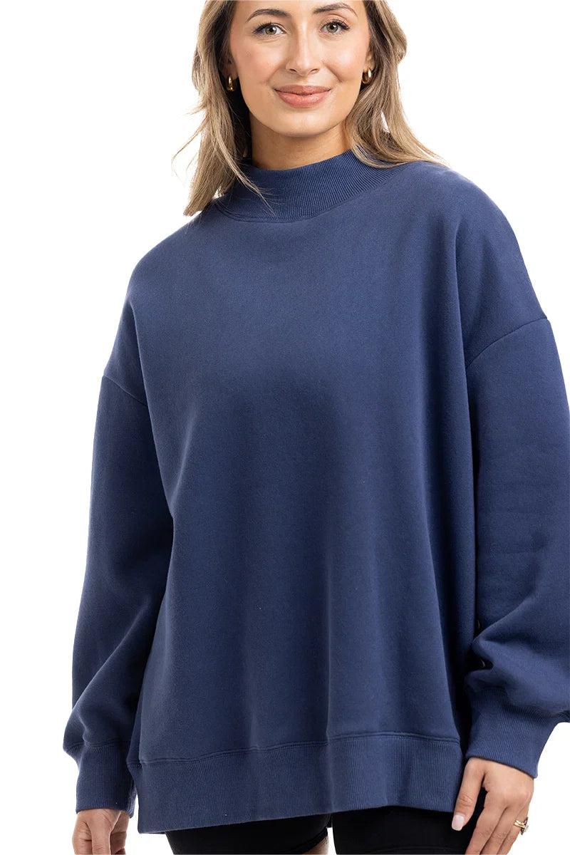 A woman with light brown hair smiles gently while wearing the Katydid Navy Mockneck Sweatshirt for women and black pants, standing against a white background.