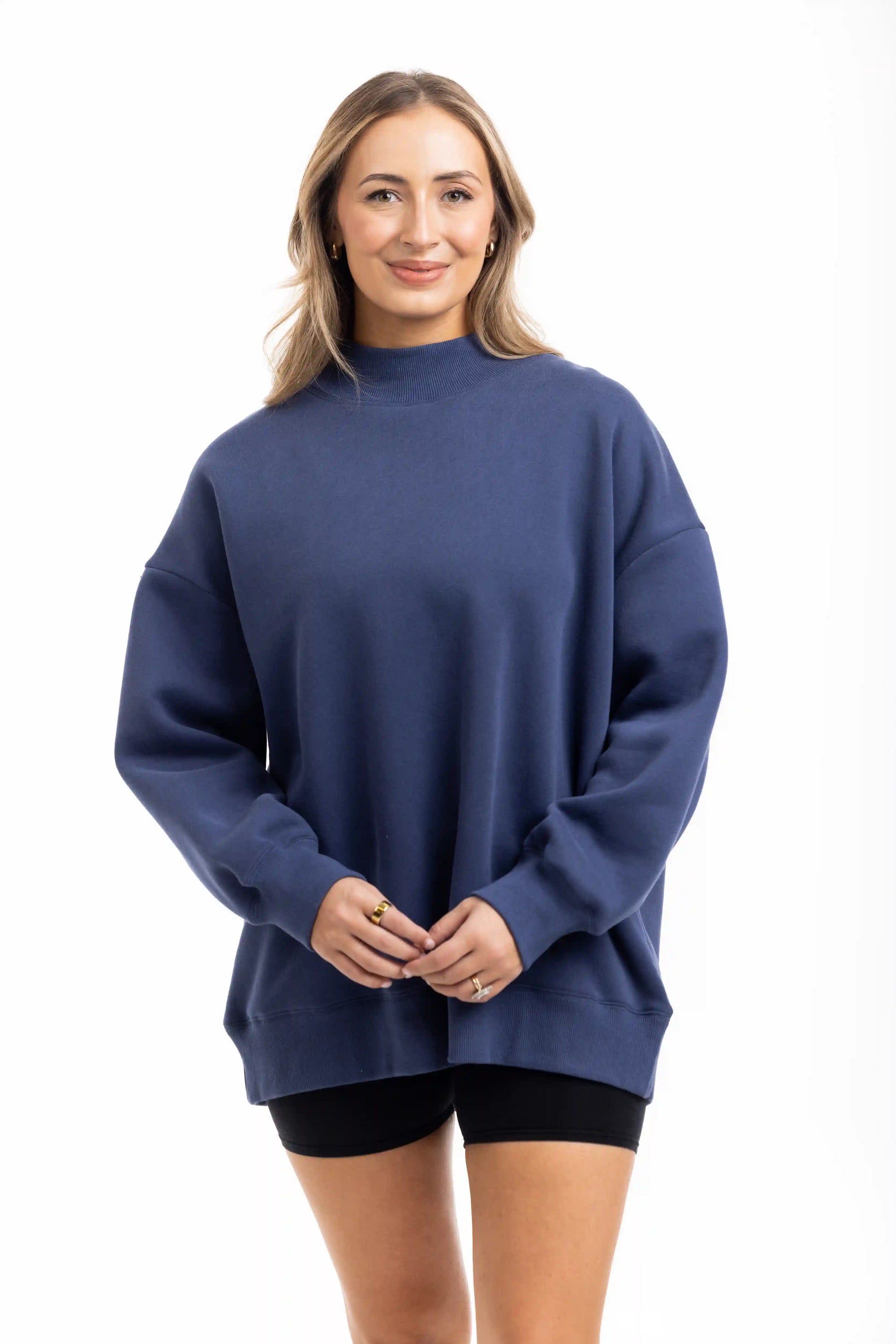 A woman with long, light brown hair smiles while wearing the Katydid Navy Mockneck Sweatshirt for women and black shorts, standing against a plain white background.