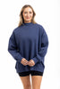 A woman with long, light brown hair smiles while wearing the Katydid Navy Mockneck Sweatshirt for women and black shorts, standing against a plain white background.