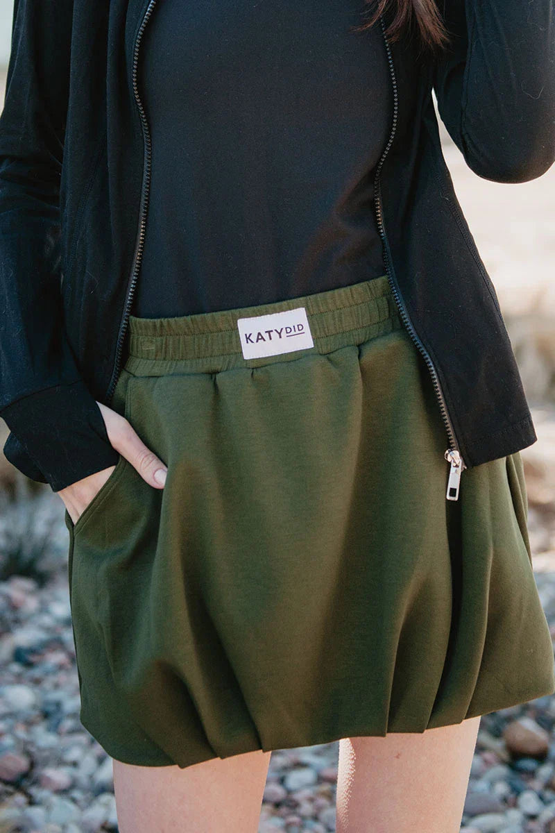 A person wears a dark long-sleeve top and the Olive Athletic Skirt for Women by Katydid, featuring soft scuba material, an elastic waistband, and a visible Katydid label. The outdoor background is blurred.