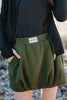 A person wears a dark long-sleeve top and the Olive Athletic Skirt for Women by Katydid, featuring soft scuba material, an elastic waistband, and a visible Katydid label. The outdoor background is blurred.