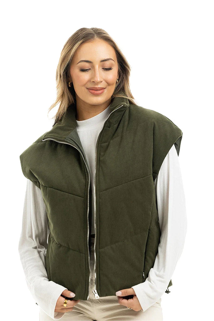 A woman with light brown hair smiles with her eyes closed, wearing the Katydid Olive Corduroy Puffer Vest over a white long-sleeve shirt, standing against a plain white background.