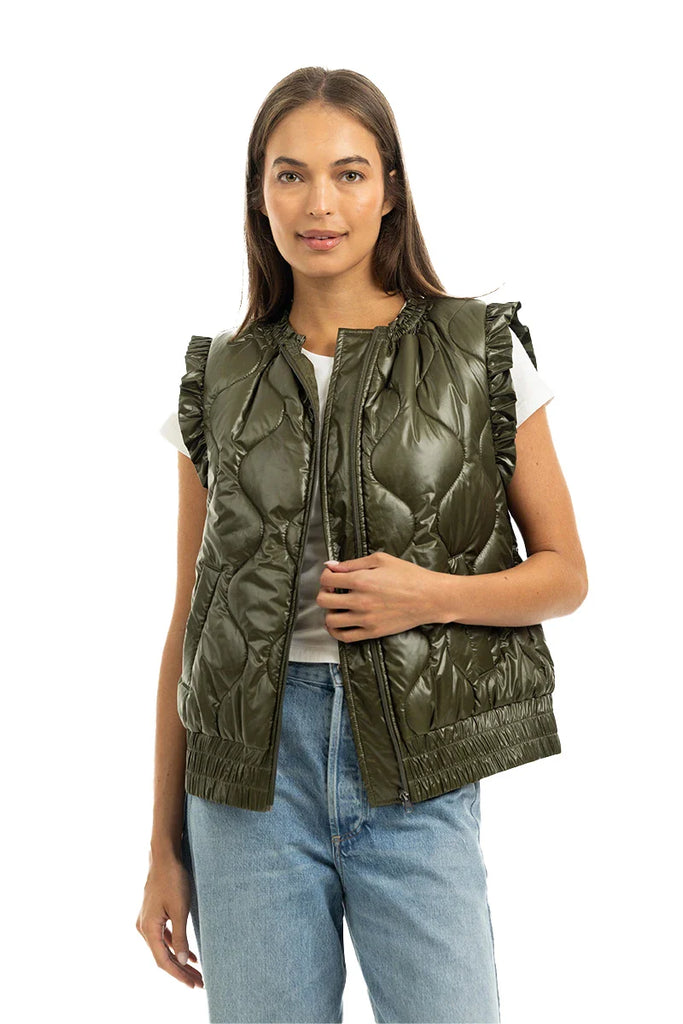 A woman wearing a white short-sleeve top, light blue jeans, and the Katydid Olive Metallic Puffer Lightweight Vest smiles against a white background.