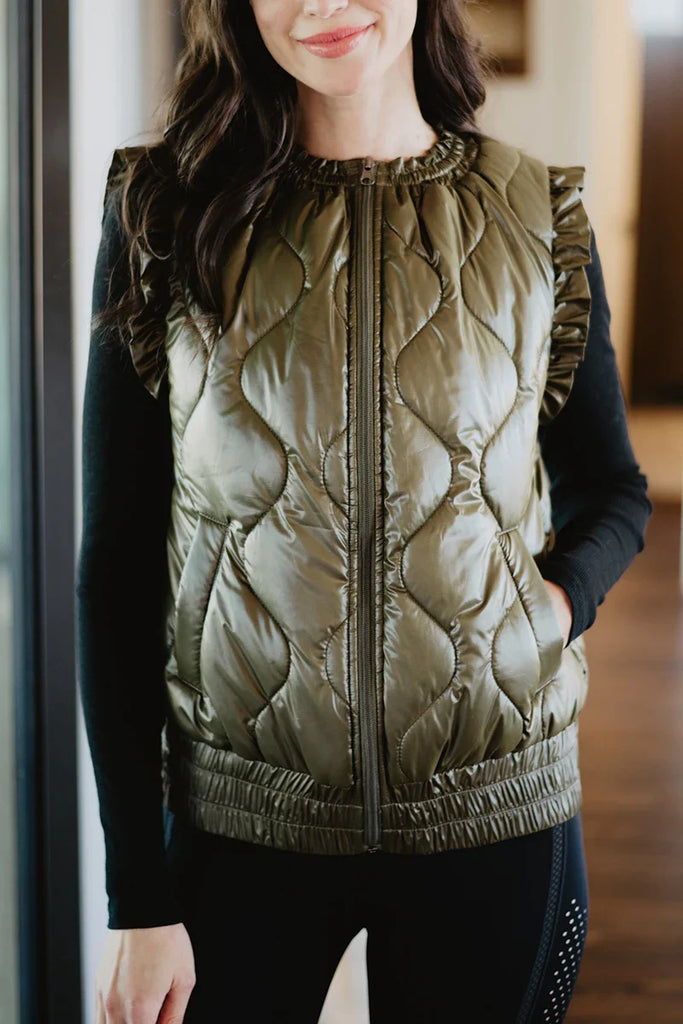 A woman stands indoors with her hands in the pockets of a Katydid Olive Metallic Puffer Lightweight Vest, worn over a black long-sleeve top.