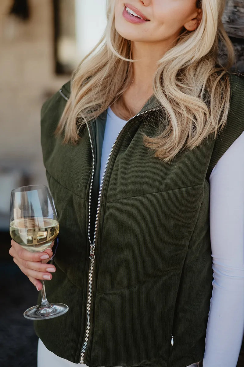 A woman with long blonde hair wears the Katydid Olive Corduroy Puffer Vest over a white shirt, holding a glass of white wine. Only her upper body and part of her face are visible.