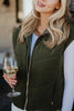 A woman with long blonde hair wears the Katydid Olive Corduroy Puffer Vest over a white shirt, holding a glass of white wine. Only her upper body and part of her face are visible.