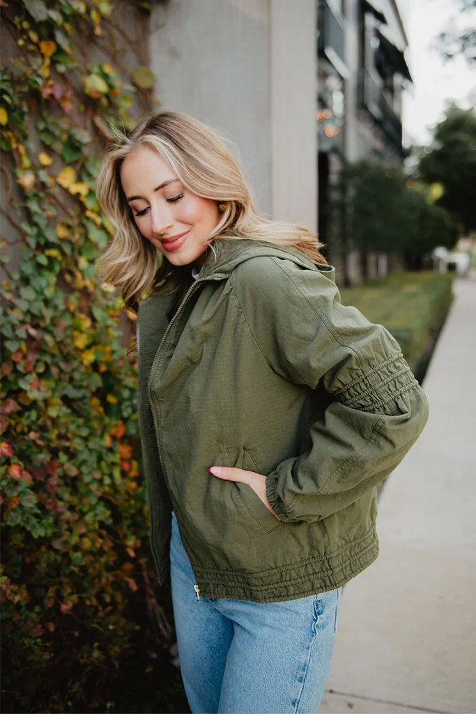 A woman with shoulder-length blonde hair, wearing the Katydid Olive Lightweight Fall Jacket and blue jeans, stands outdoors near a wall of green and red ivy, smiling with eyes closed and one hand in her pocket.