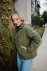 A woman with shoulder-length blonde hair, wearing the Katydid Olive Lightweight Fall Jacket and blue jeans, stands outdoors near a wall of green and red ivy, smiling with eyes closed and one hand in her pocket.
