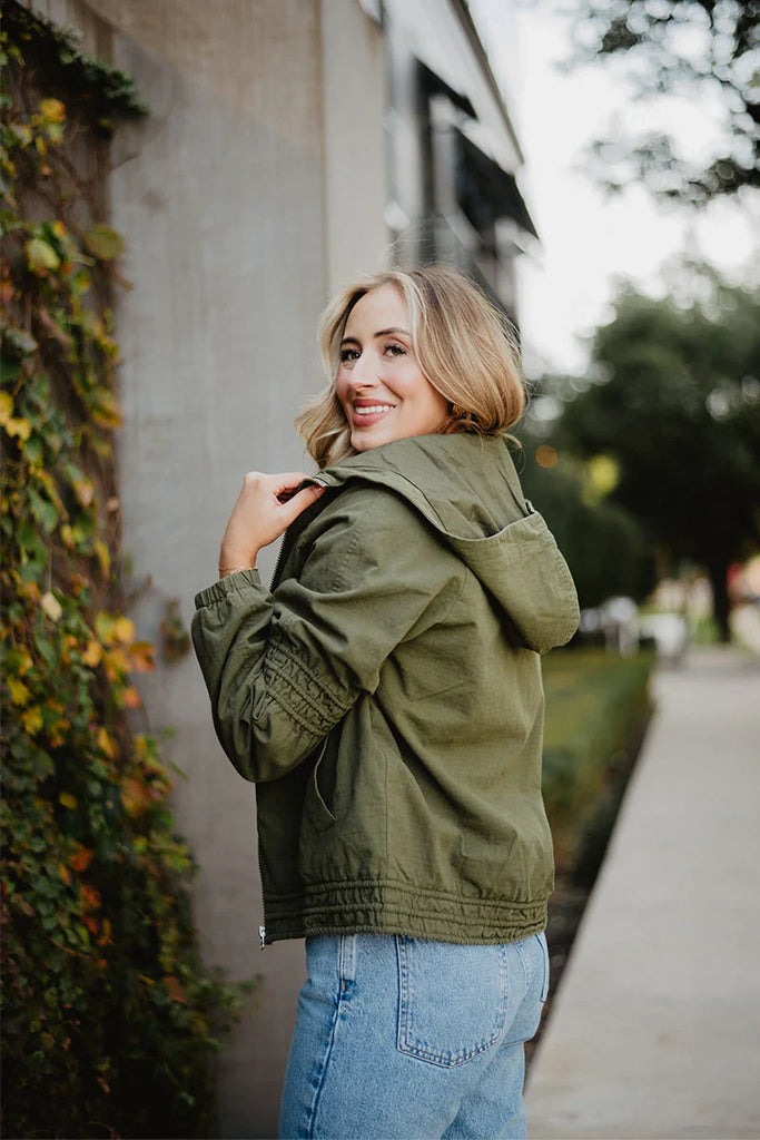 A woman with blonde hair, wearing the Katydid Olive Lightweight Fall Jacket for women and blue jeans, stands on a sidewalk beside greenery and a building wall, looking back over her shoulder with a smile.