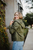 A woman with blonde hair, wearing the Katydid Olive Lightweight Fall Jacket for women and blue jeans, stands on a sidewalk beside greenery and a building wall, looking back over her shoulder with a smile.