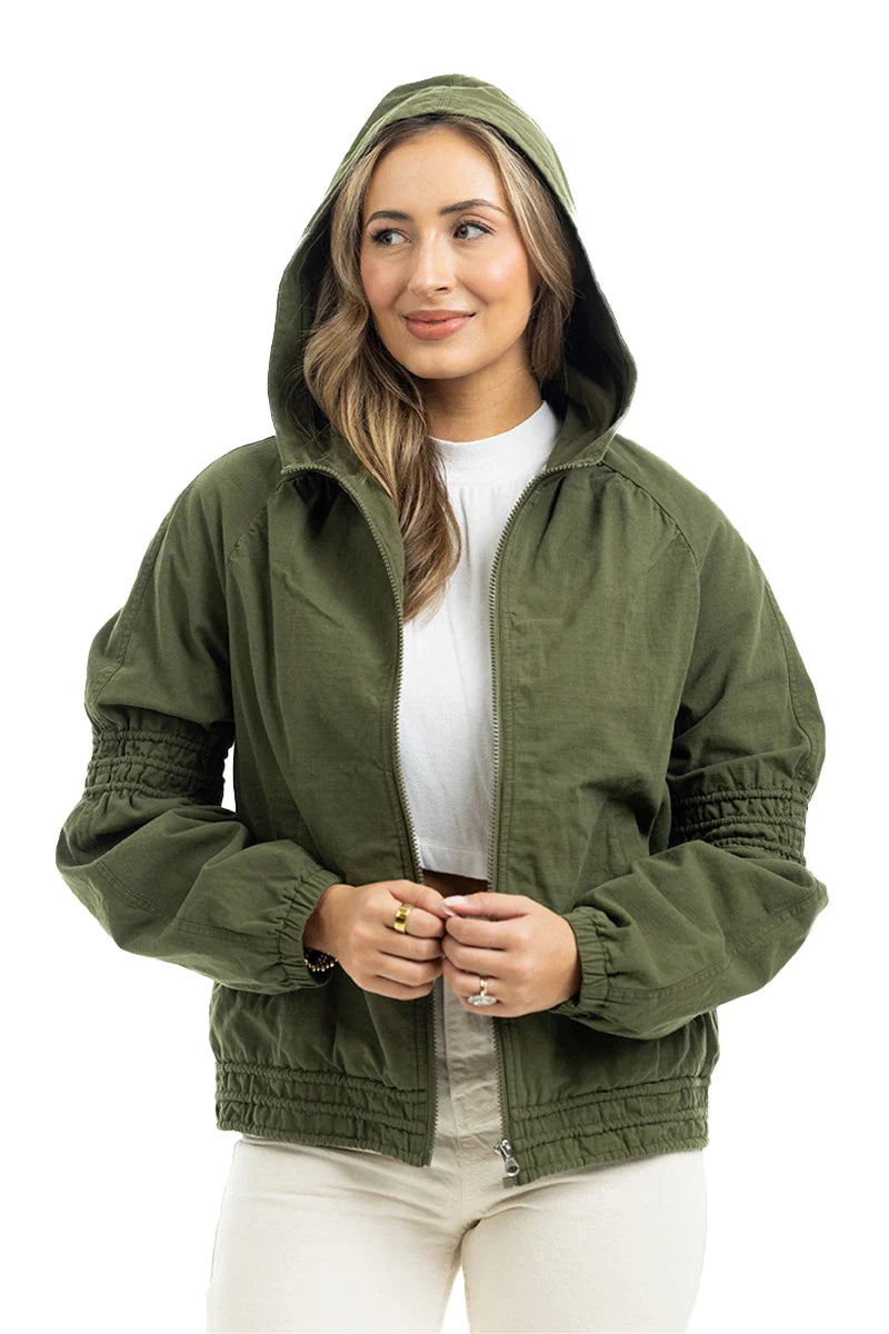 A woman models the Katydid Olive Lightweight Fall Jacket for women, wearing it over a white top and light pants. She stands facing forward, gently smiling with her hands on the zipper against a plain white background.