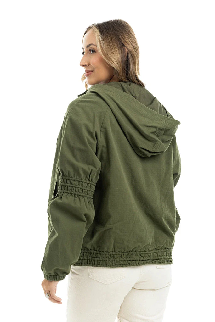 A woman with light brown hair stands against a white background, wearing the Katydid Olive Lightweight Fall Jacket Women and white pants, looking over her shoulder.