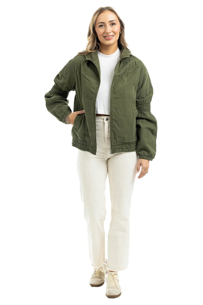 A woman smiles and stands against a plain white background, wearing the Katydid Olive Lightweight Fall Jacket over a white shirt, paired with cream pants and beige sneakers.