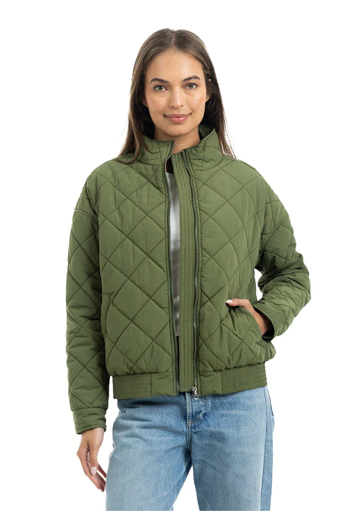 A woman smiles slightly, standing with one hand in her pocket, wearing the Katydid Olive Sporty Diamond Quilted Jacket and blue jeans.